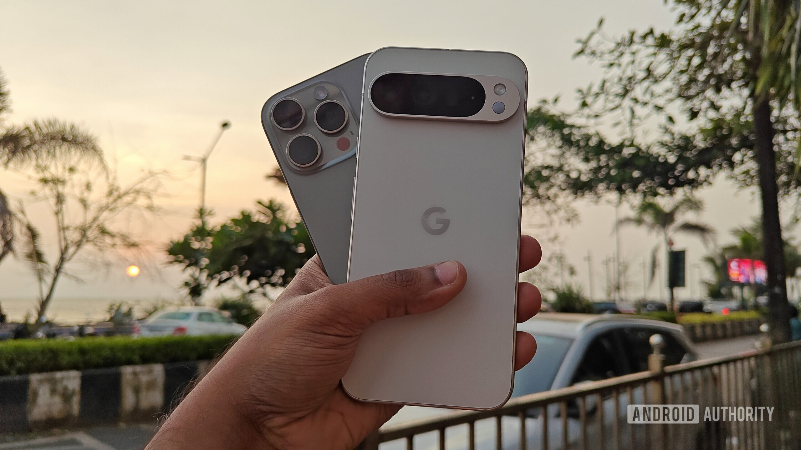 Apple iPhone 16 Pro and Google Pixel 9 Pro XL Apple iPhone 16 Pro and Google Pixel 9 Pro XL held out in hand, against the backdrop of a sunset on an urban promenade